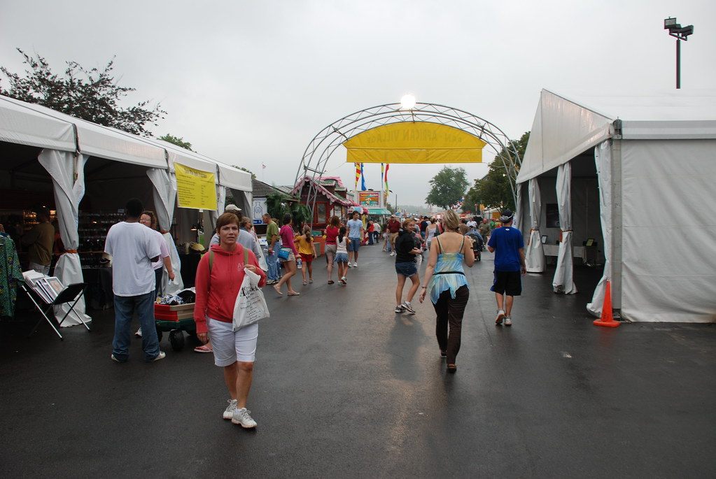 Visitors walking between white vendor tents under an arched entry sign on the fairgrounds on a wet day