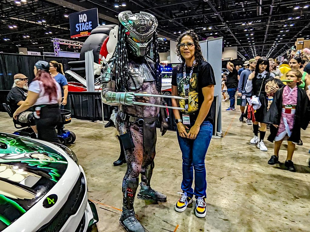 A Predator cosplayer with wrist blades posing with a fan on the MegaCon Orlando 2023 show floor, with the HQ Stage sign and crowds behind