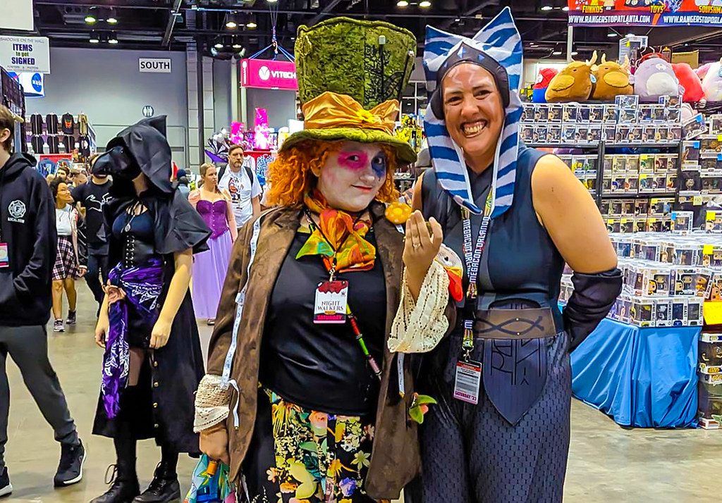 Two smiling cosplayers, one as the Mad Hatter, beside a toy vendor's booth on the MegaCon Orlando 2023 show floor