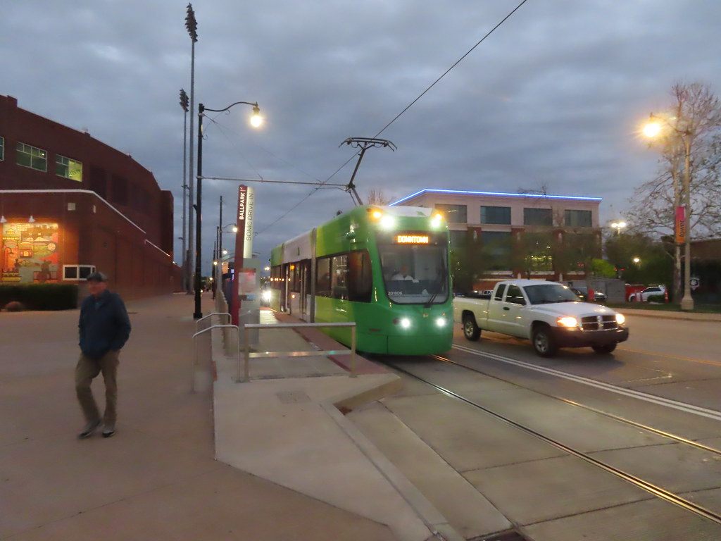 A green Oklahoma City streetcar pulling into the Ball Park stop at dusk as a man walks along the platform