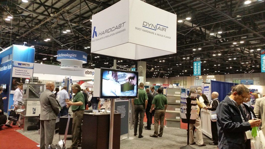 Exhibitors and visitors talking at HVAC product booths under a hanging brand sign on the AHR Expo show floor in Orlando