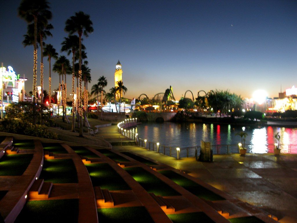 Palm trees wrapped in lights along a lagoon promenade at dusk, with a clock tower and lit restaurants on the far side