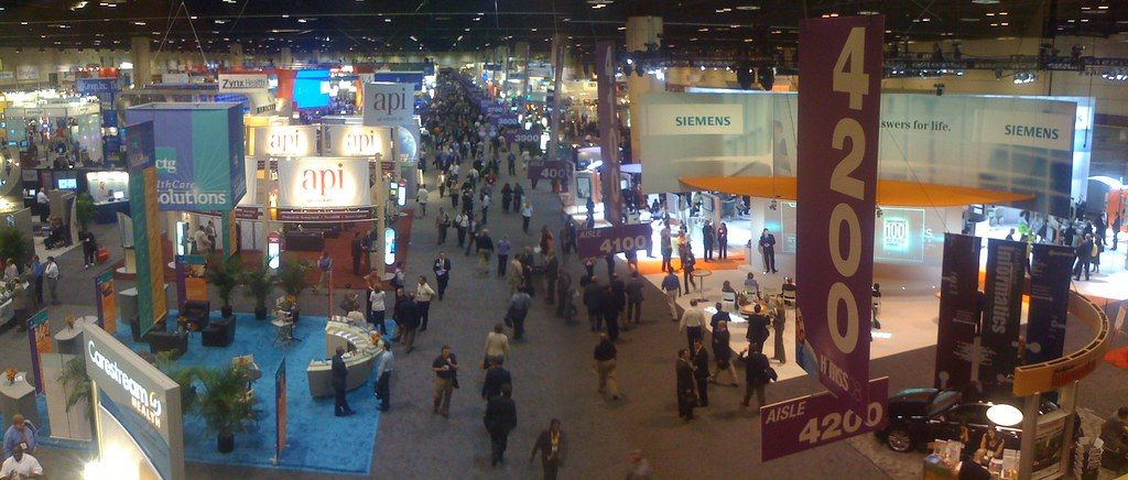 A long aisle of the HIMSS08 vendor expo in Orlando seen from above, with large exhibitor booths, hanging aisle signs and attendees walking between them