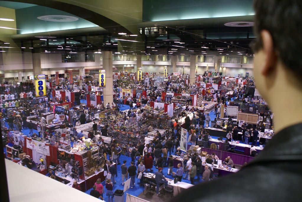 A packed comic-convention show floor seen from a balcony, with rows of vendor booths, banners and crowds in the aisles
