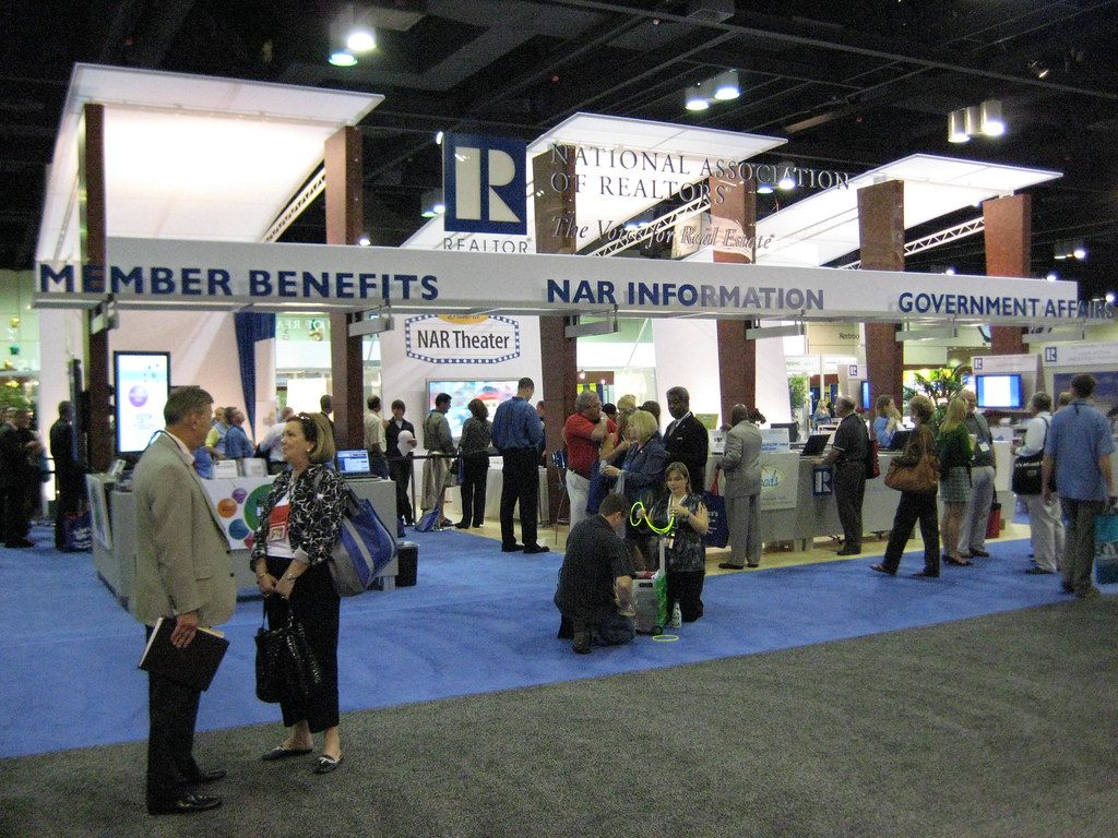 Attendees with badges and tote bags at the National Association of Realtors member-services booth on a blue-carpeted expo floor