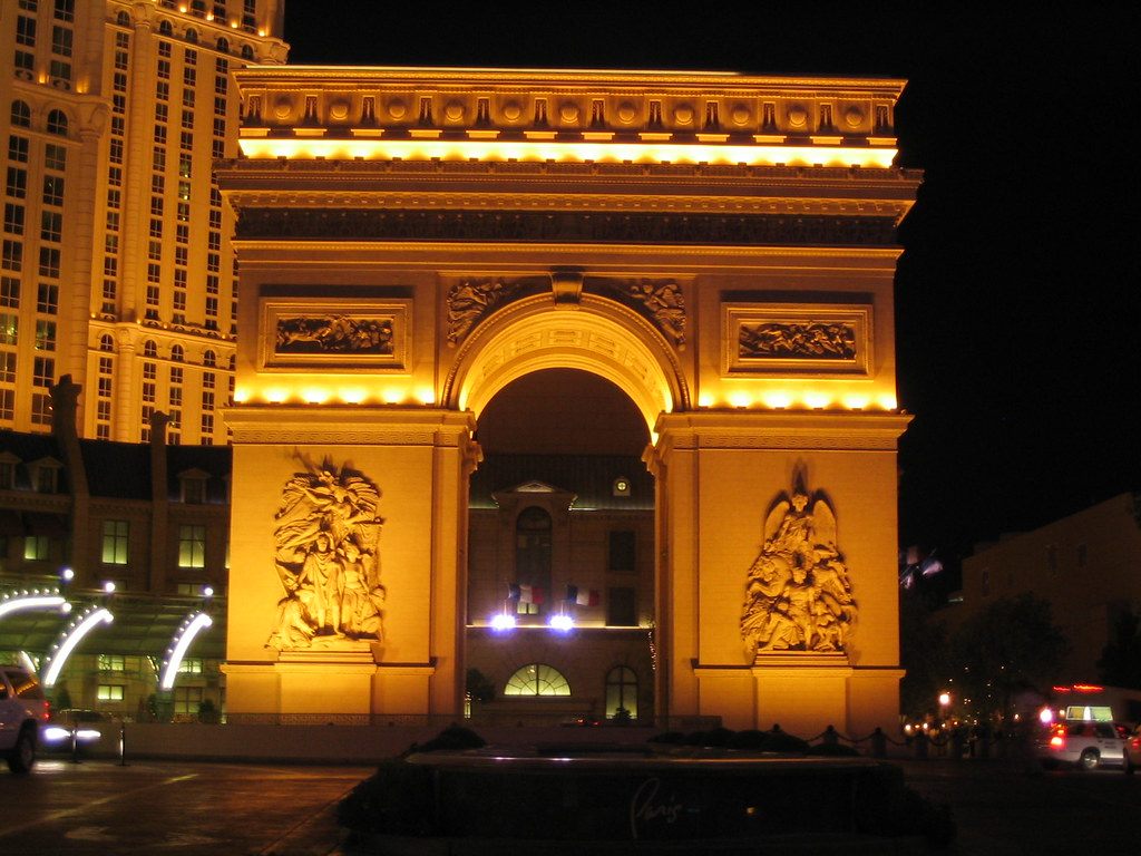 The two-thirds-size Arc de Triomphe replica lit gold at night in front of Paris Las Vegas, with the hotel tower at left