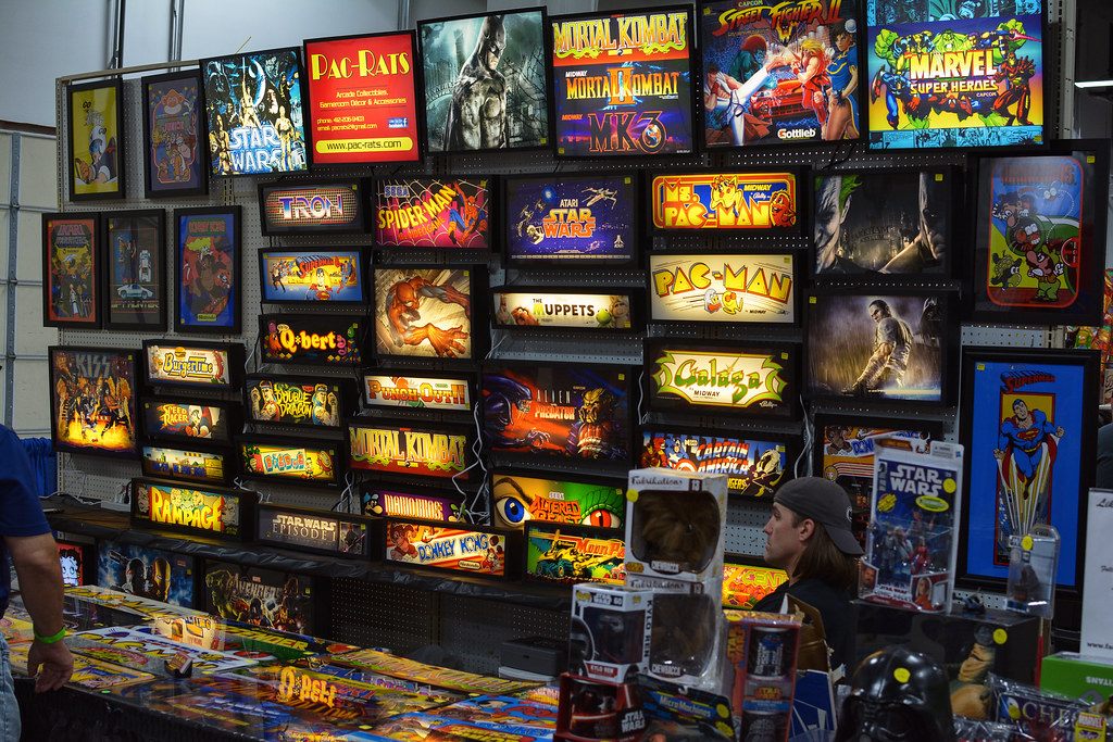 A wall of lit arcade marquees, Pac-Man to Mortal Kombat, above a vendor table at Retro Con in Oaks, PA