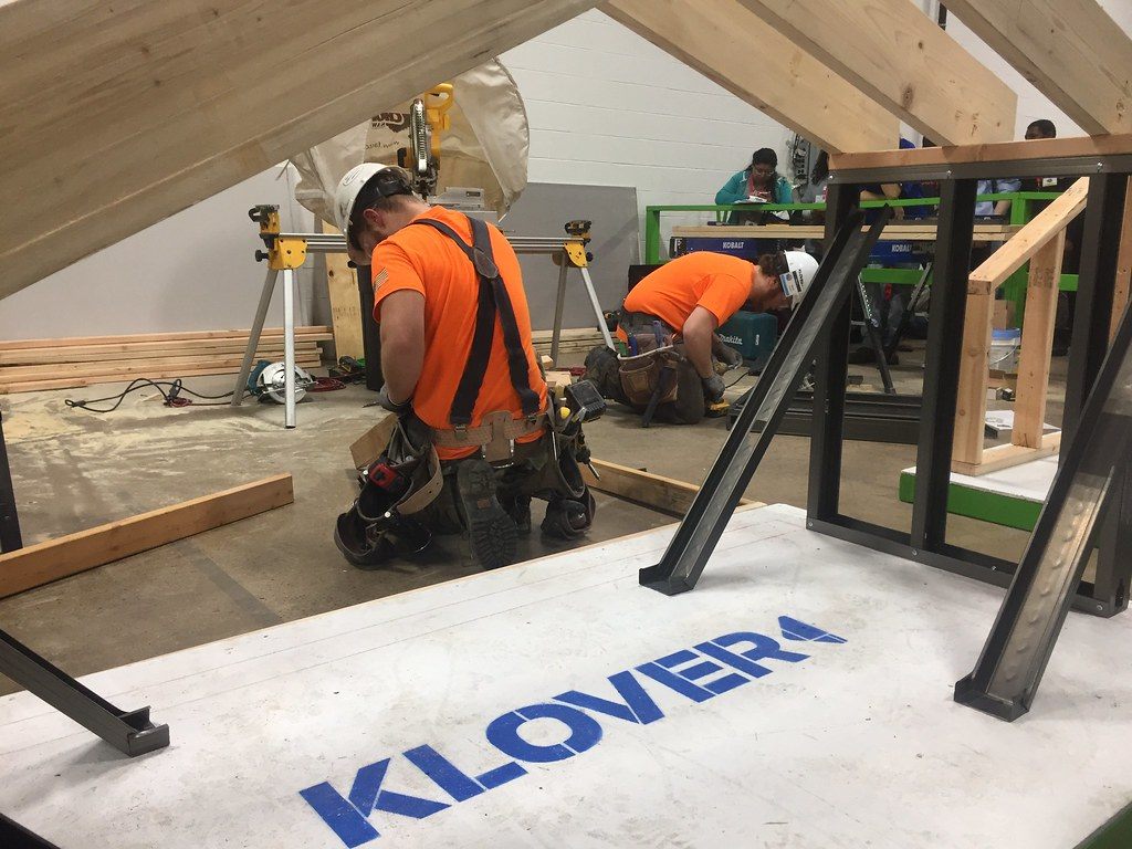 Two carpenters in hard hats and orange shirts building a timber roof frame on a concrete exhibit floor at the ManuFest manufacturing event in Oaks, Pennsylvania