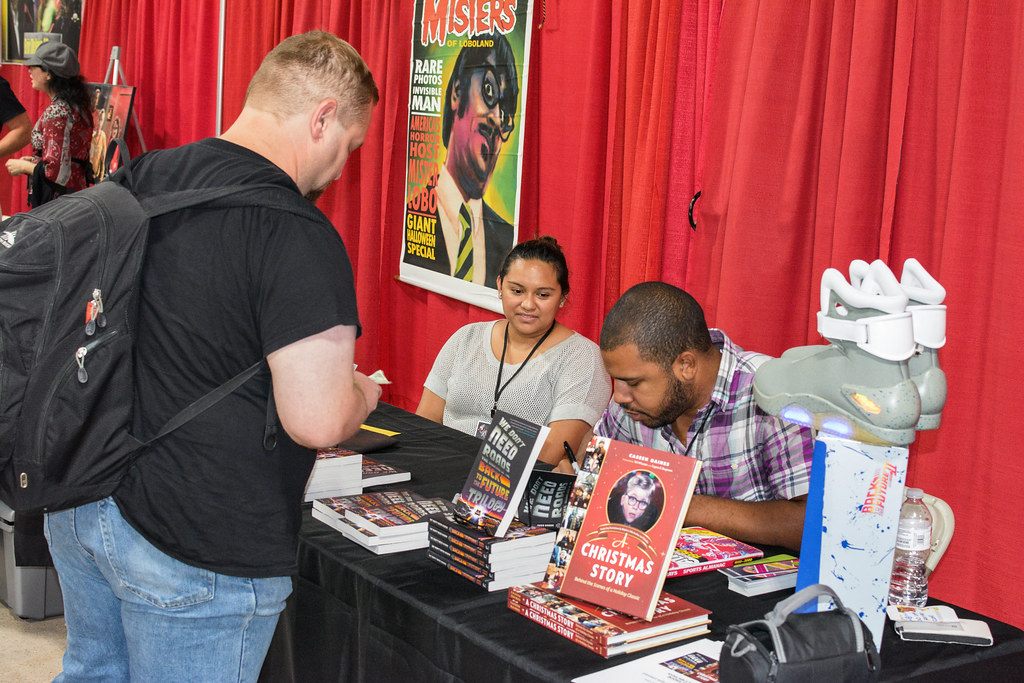A visitor with a backpack chats with two authors signing books at a red-draped table during Retro Con in Oaks, PA