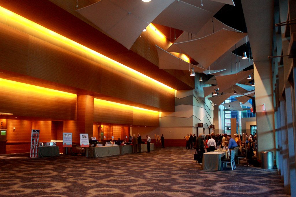 Attendees mingle at tables and registration desks in a warmly lit Phoenix Convention Center lobby with sail-like ceiling panels