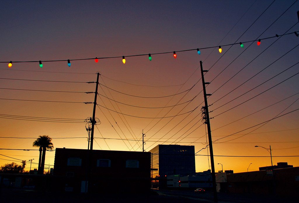Strings of colored bulbs over Roosevelt Street at sunset, with low buildings and a palm tree in silhouette