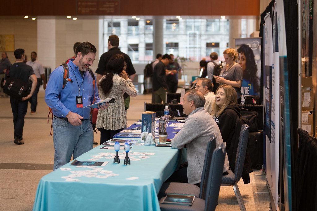 An attendee with a backpack chats with exhibitors seated at a blue-draped Intel table in the Raleigh Convention Center lobby during All Things Open 2017