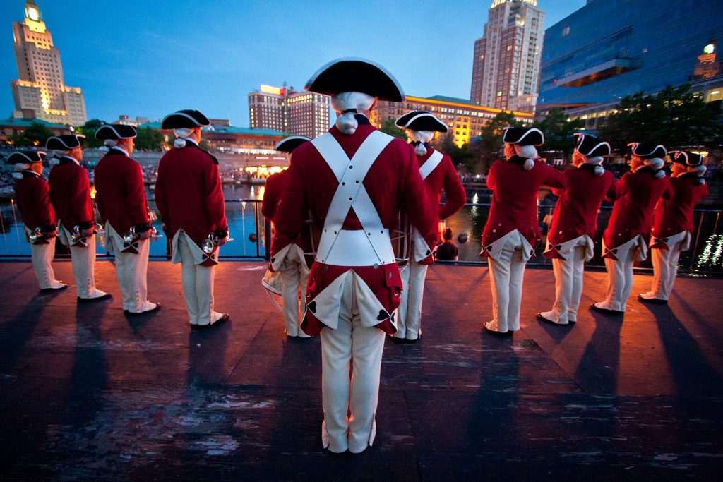 The U.S. Army Old Guard Fife and Drum Corps in red coats and tricorn hats stand on a riverside stage at dusk during WaterFire in Providence