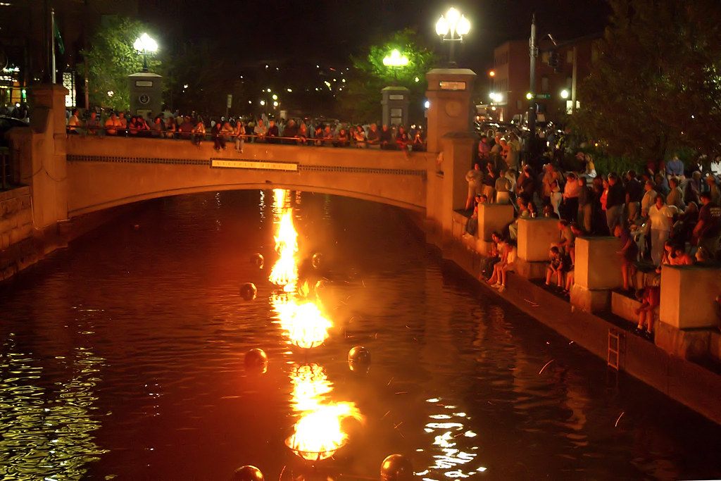 Crowds line a footbridge and the riverwalk steps as WaterFire braziers blaze on the river in downtown Providence at night