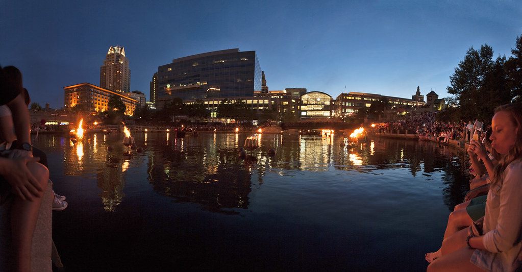 Panoramic dusk view of WaterFire with spectators seated along the river basin and the downtown Providence skyline lit behind