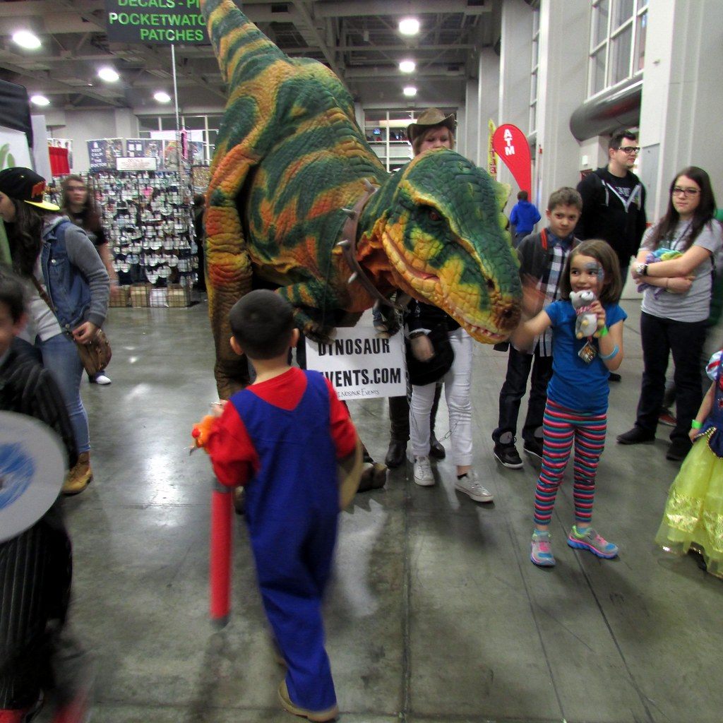 A costumed raptor greets kids in cosplay in a wide aisle at FanX Salt Lake 2015
