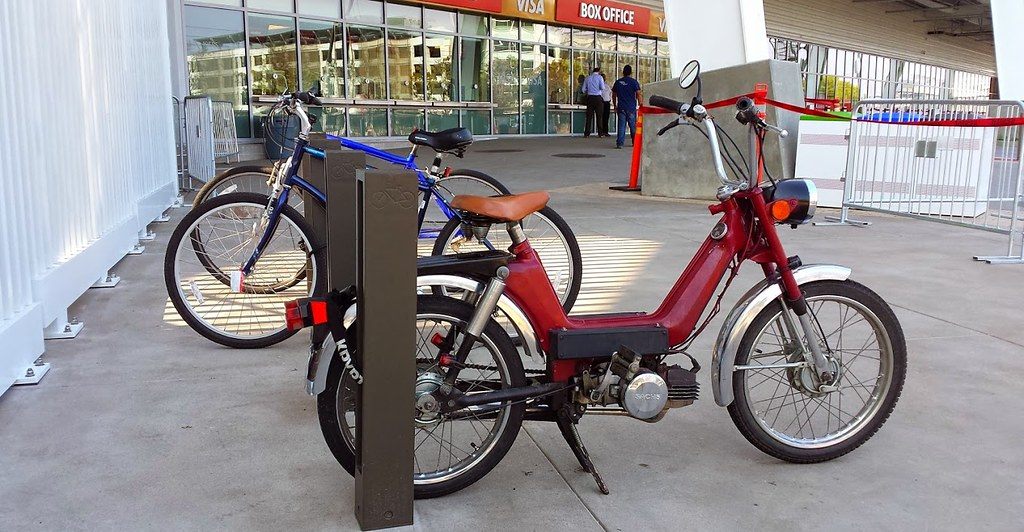 Bicycles and a red moped locked at a bike rack beside the glass box office at Levi's Stadium