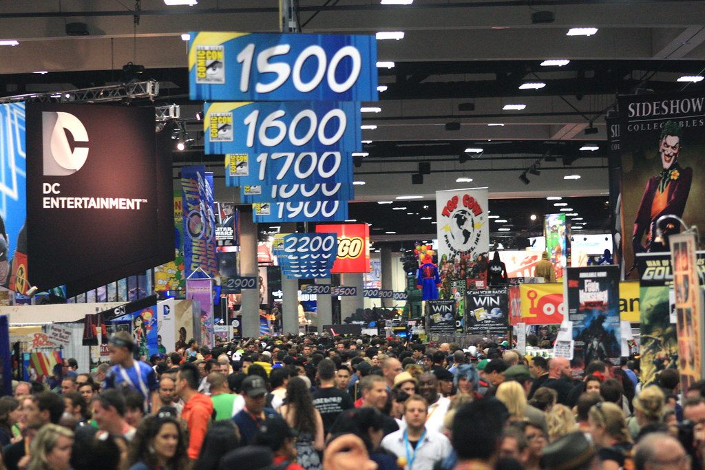 A packed San Diego Comic-Con exhibit hall with hanging aisle-number signs and crowds between giant publisher booths