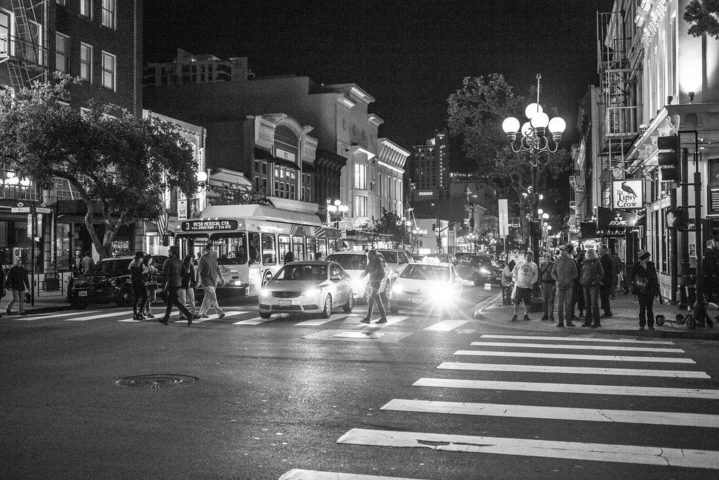 Black-and-white night scene of people crossing a Gaslamp Quarter street past a trolley bus and old brick buildings