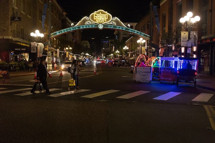 People crossing Fifth Avenue at night under the lit Gaslamp Quarter arch, with pedicabs glowing at the curb