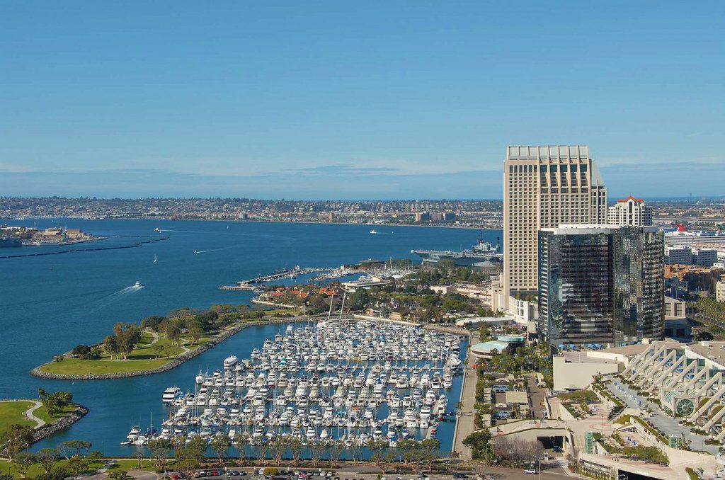 Aerial view of a downtown San Diego marina packed with white boats, a park peninsula curving around it, glass hotel towers at right and the bay beyond