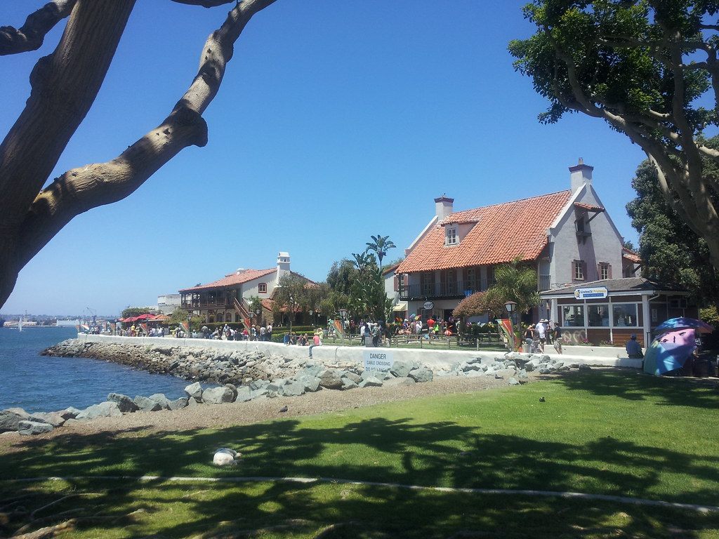 Visitors strolling the bayside lawn and paths of Seaport Village under red-tiled roofs on a sunny day