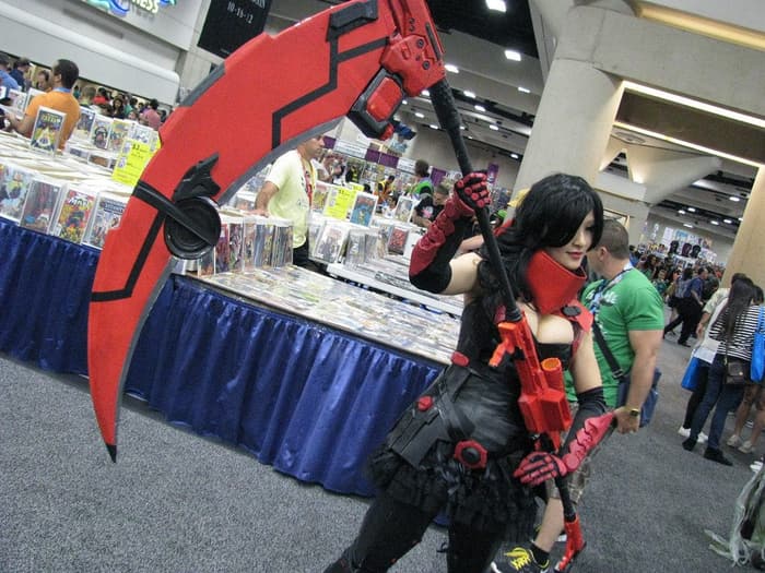 A cosplayer with a giant red scythe walking past comic-book dealer tables in a San Diego Convention Center exhibit hall at Comic-Con 2012