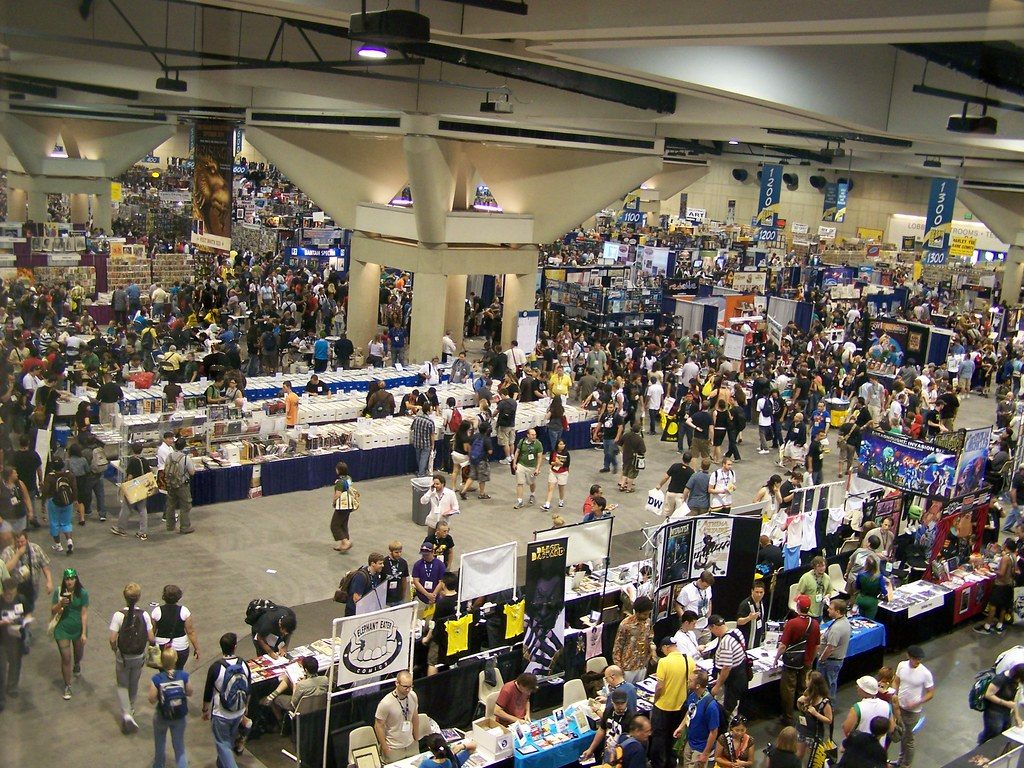 Overhead view of a packed San Diego Convention Center exhibit hall during Comic-Con 2008, with booth rows, aisle number banners and concrete ceiling beams