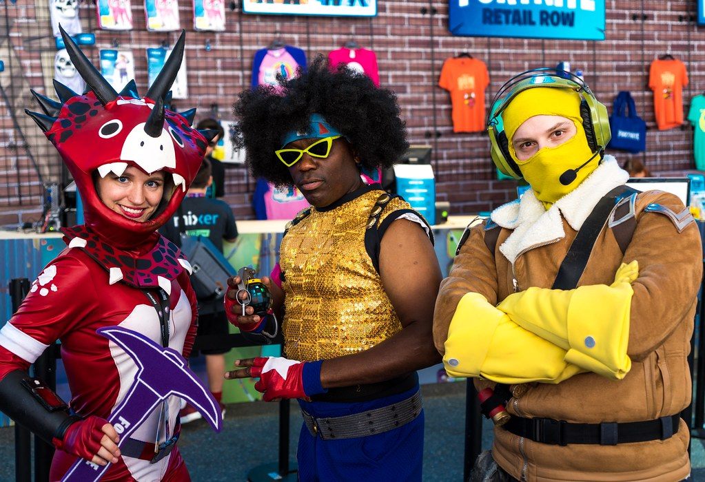 Three Fortnite cosplayers posing in front of a merchandise wall at PAX West 2018 in Seattle