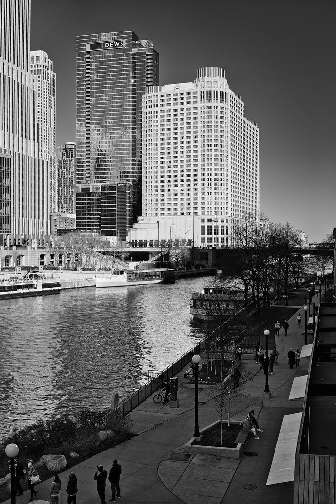 Black-and-white view east along the Chicago River and its riverwalk toward the Sheraton Grand Chicago and the Loews tower, with tour boats moored