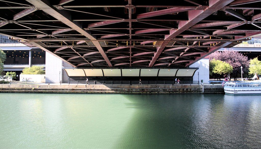 Looking out from under a red steel bridge across the green Chicago River to a riverside walkway and a tour boat