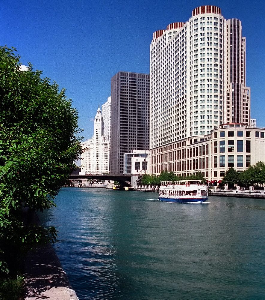The Sheraton hotel tower and its low riverfront wing on the north bank of the Chicago River, with a tour boat passing and the Wrigley Building in the distance
