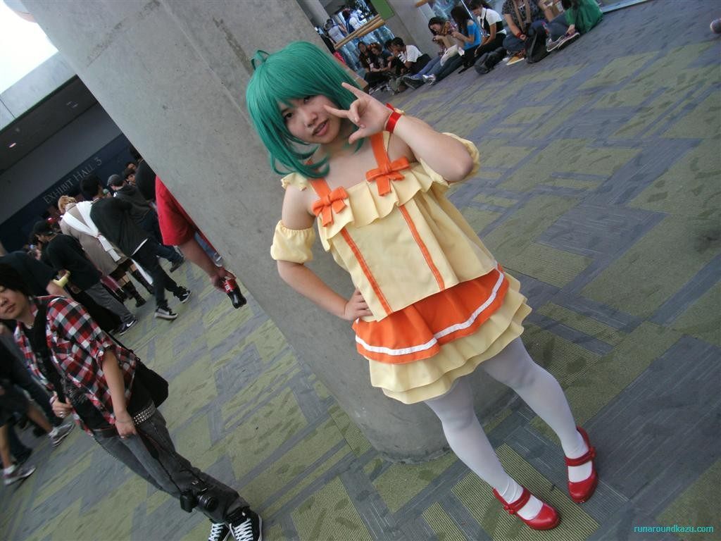 A costumed attendee in a yellow and orange dress posing under the convention center's concrete columns as crowds sit on the carpet behind during FanimeCon 2010