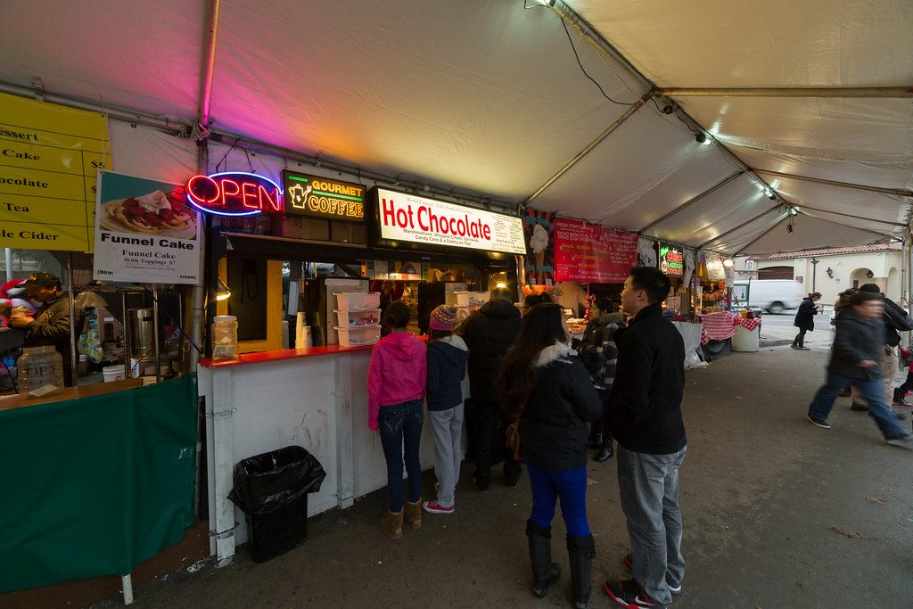 Visitors in winter coats lining up at hot chocolate and funnel cake stands under a festival tent in Plaza de César Chávez