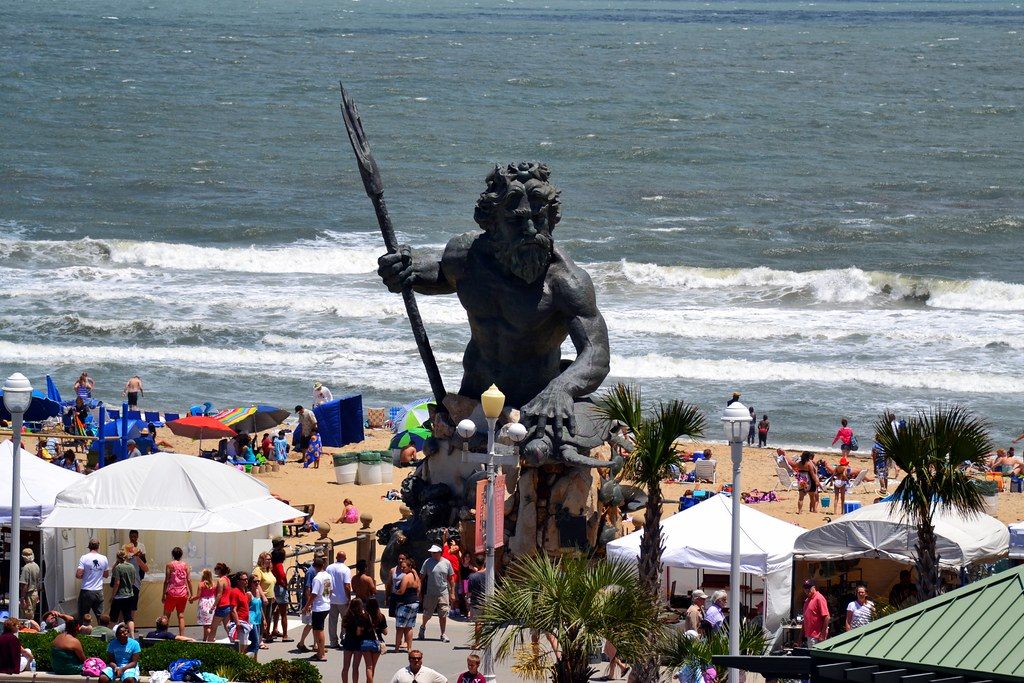 Crowds and white vendor tents around the King Neptune statue on the Virginia Beach boardwalk during the Boardwalk Art Show, with surf behind