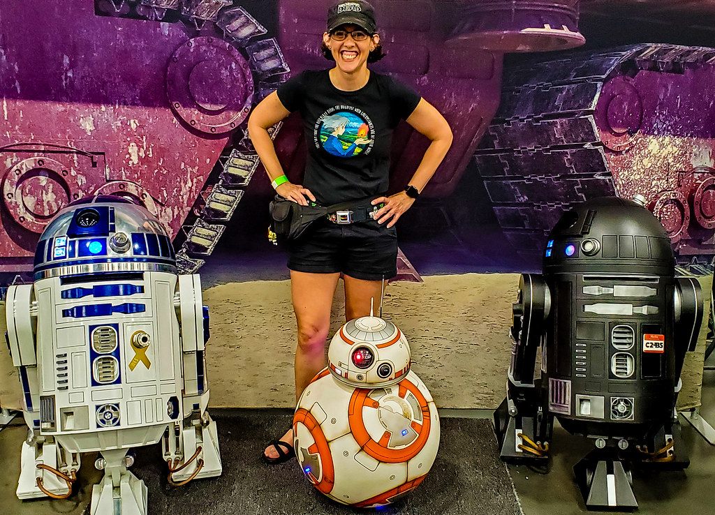 A smiling fan in a black cap poses between R2-D2, BB-8 and a black astromech droid in front of a Star Wars backdrop at Tidewater Comicon 2019