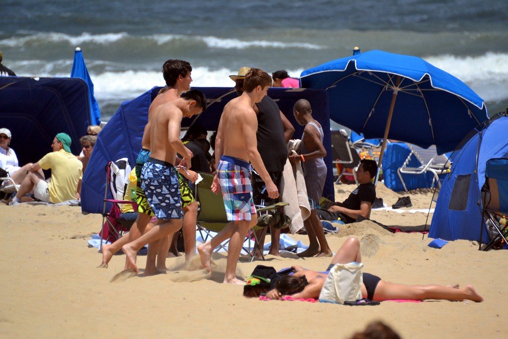 Beachgoers walking between blue umbrellas and tents on a packed Virginia Beach oceanfront in summer