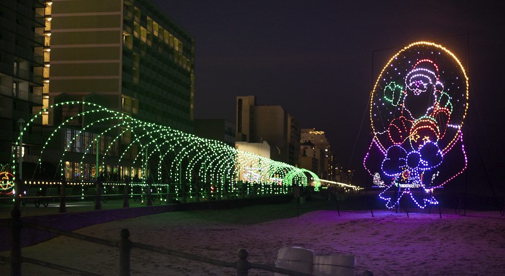 A tunnel of green and white light arches and a lit Santa display on the sand beside the Virginia Beach boardwalk hotels at night