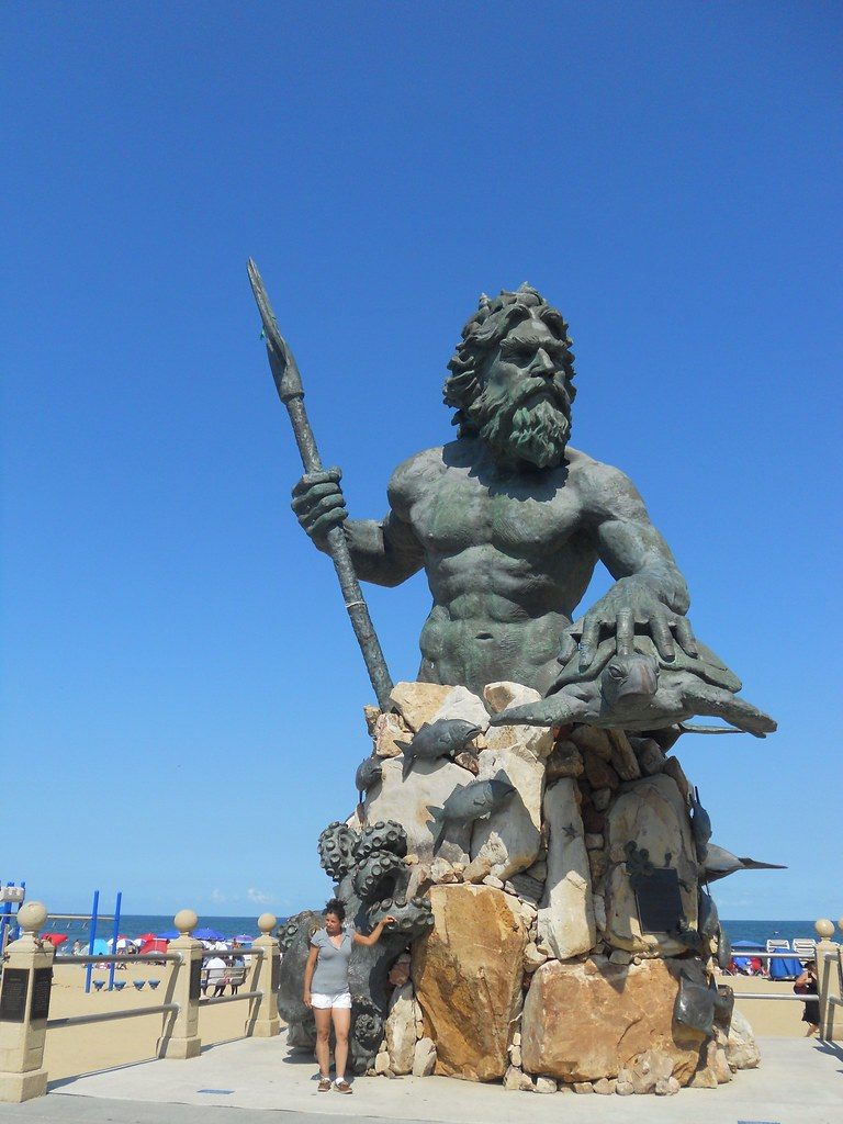 The bronze King Neptune statue holding a trident above a rock base on the Virginia Beach boardwalk, with beach umbrellas behind