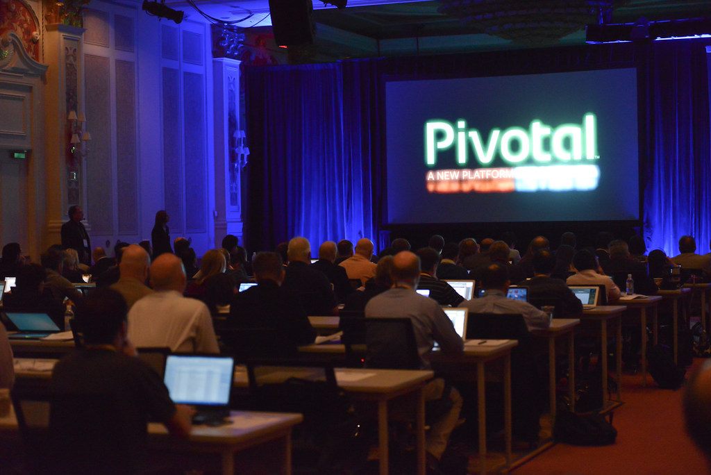 Attendees with laptops at long tables in a darkened Venetian meeting room facing a big screen during EMC World 2013