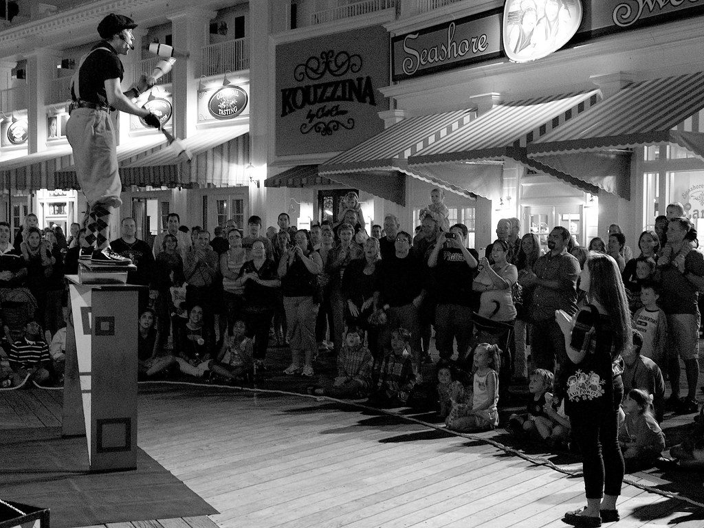 A crowd of families on Disney's BoardWalk at night watching a street performer juggle on a raised box in front of the shopfronts