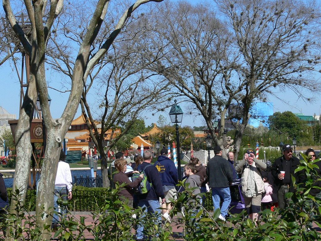 Visitors strolling a tree-lined World Showcase walkway at Epcot on a sunny day, with pagoda roofs behind