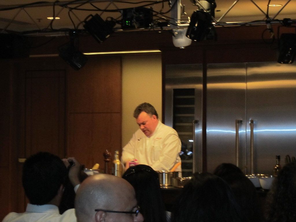 A chef in whites cooks on a demo stage under rigged lights while guests watch at the Cadillac and Westchester Magazine Wine & Food Weekend