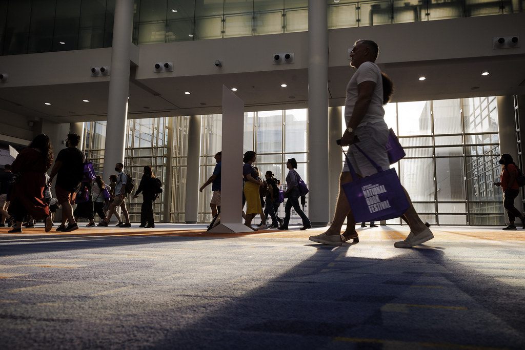 Silhouetted festival-goers carrying tote bags cross the sunlit glass lobby of the convention center during the National Book Festival