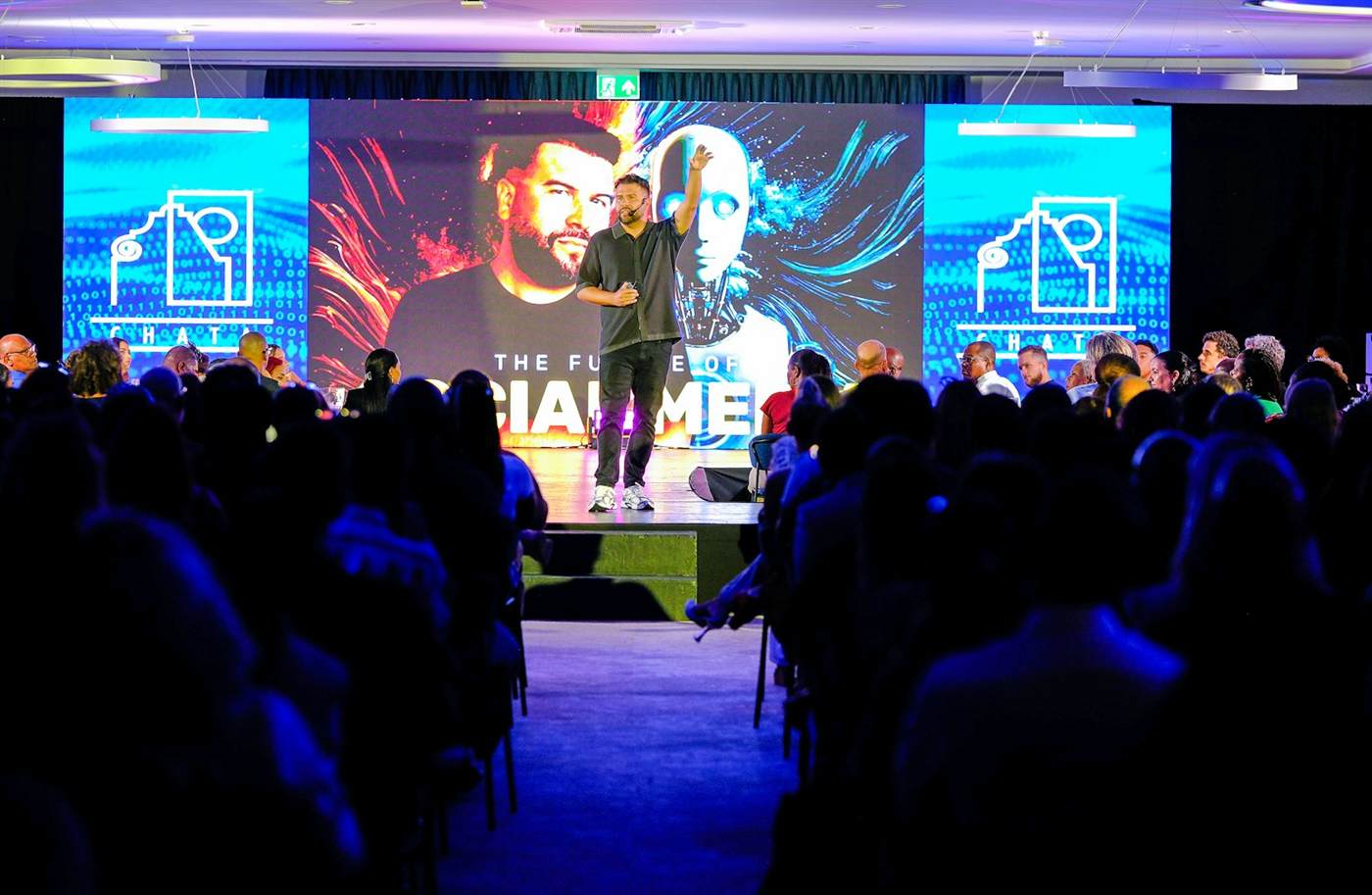 A presenter on stage in front of LED walls with a seated audience lit in blue