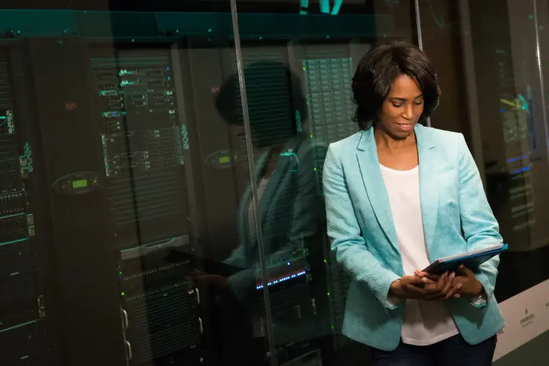 Woman in a teal blazer reviewing a tablet beside glass-fronted server racks