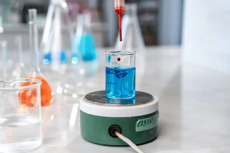A dropper adds red liquid to a blue solution in glassware on a magnetic stirrer
