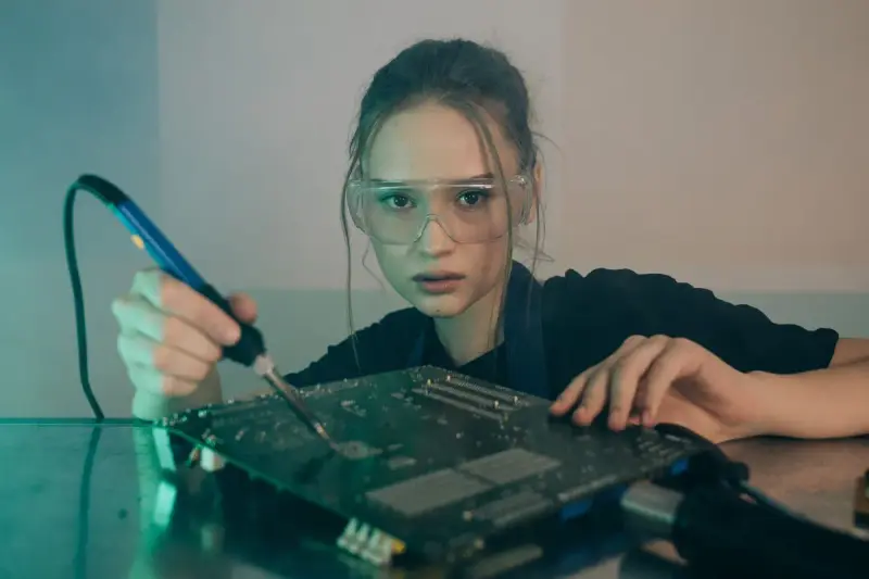 A young woman in safety glasses solders a circuit board