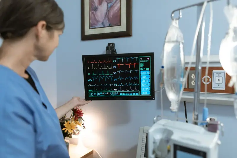Nurse in blue scrubs examining a bedside monitor showing vital signs in a hospital room