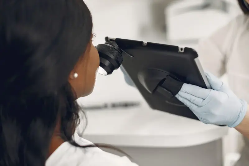 Gloved hand holds a tablet fitted with a dermatoscope lens close to a patient's cheek